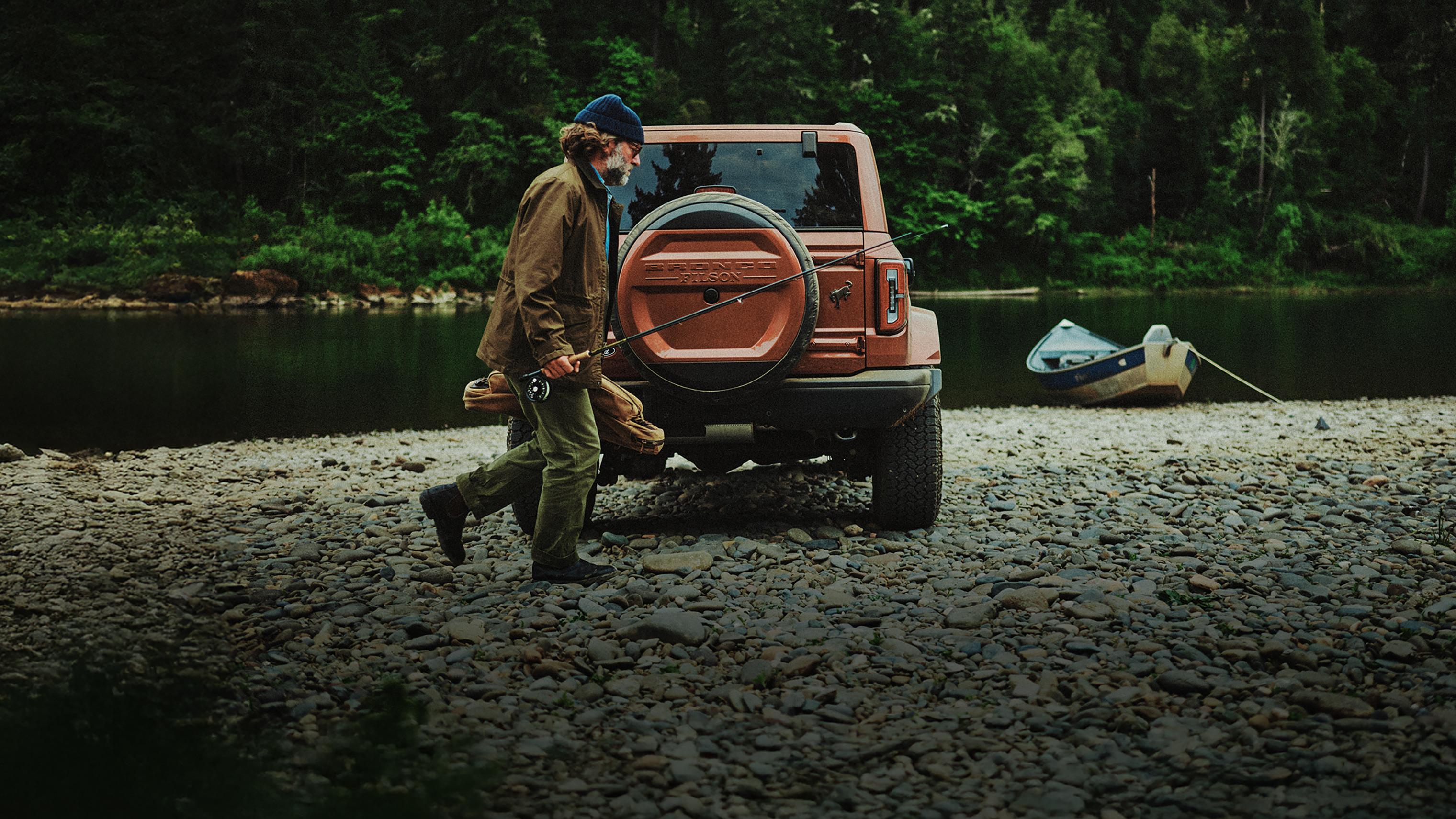 A man carrying a fishing pole and a storage bag near a 2027 Ford Bronco® Filson SUV parked on a rocky lakeshore