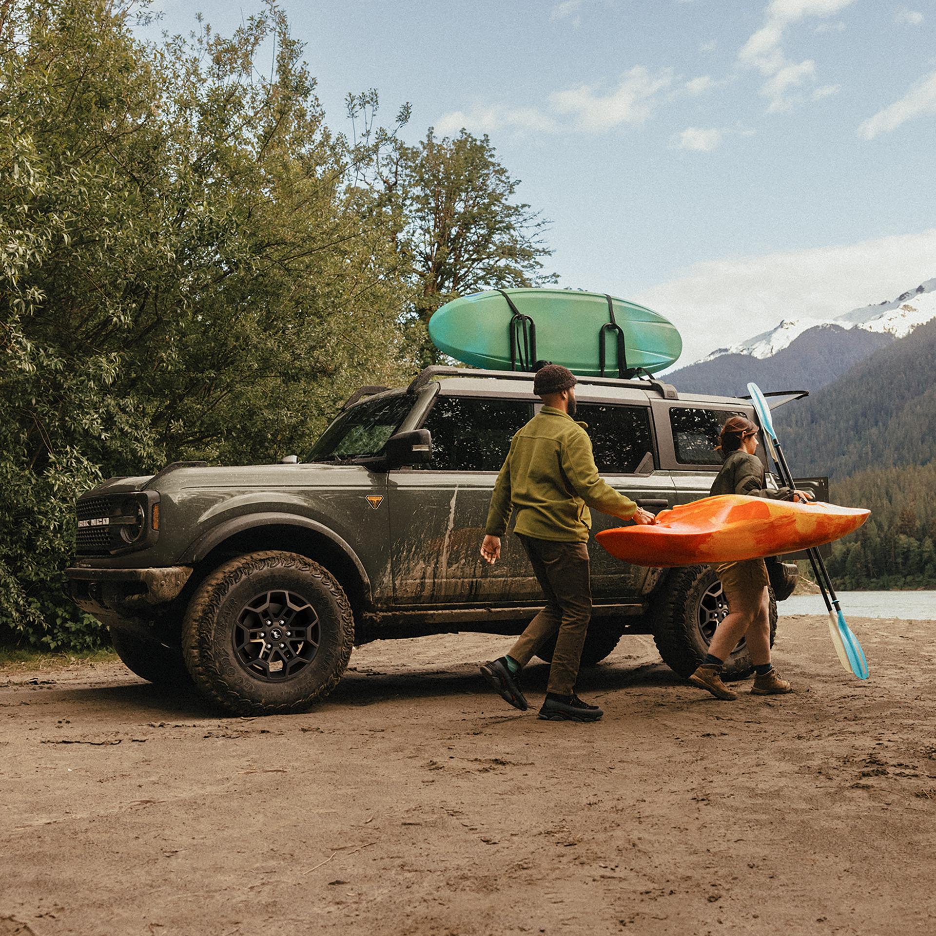 A man and woman carrying a kayak and paddle away from a 2026 Ford Bronco® SUV parked on a mud flat