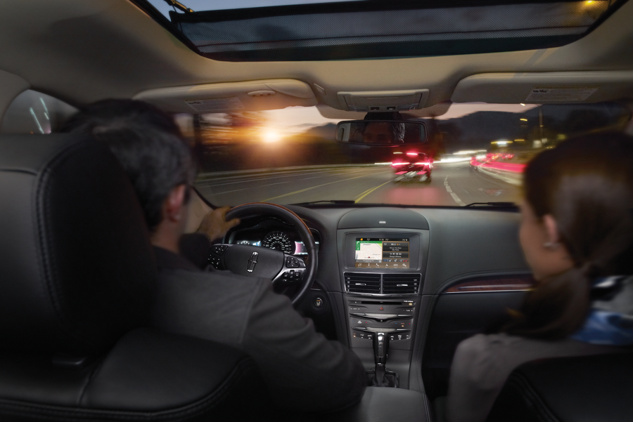 A couple is seen driving a Lincoln M K T while brake lights of the car in front of them are seen through the windshield.