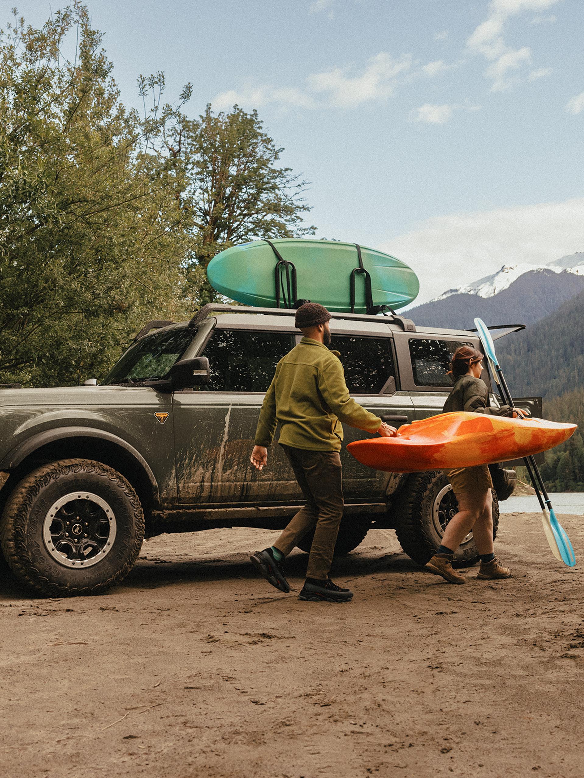 A man and woman carrying a kayak and paddle away from a 2026 Ford Bronco® SUV parked on a mud flat