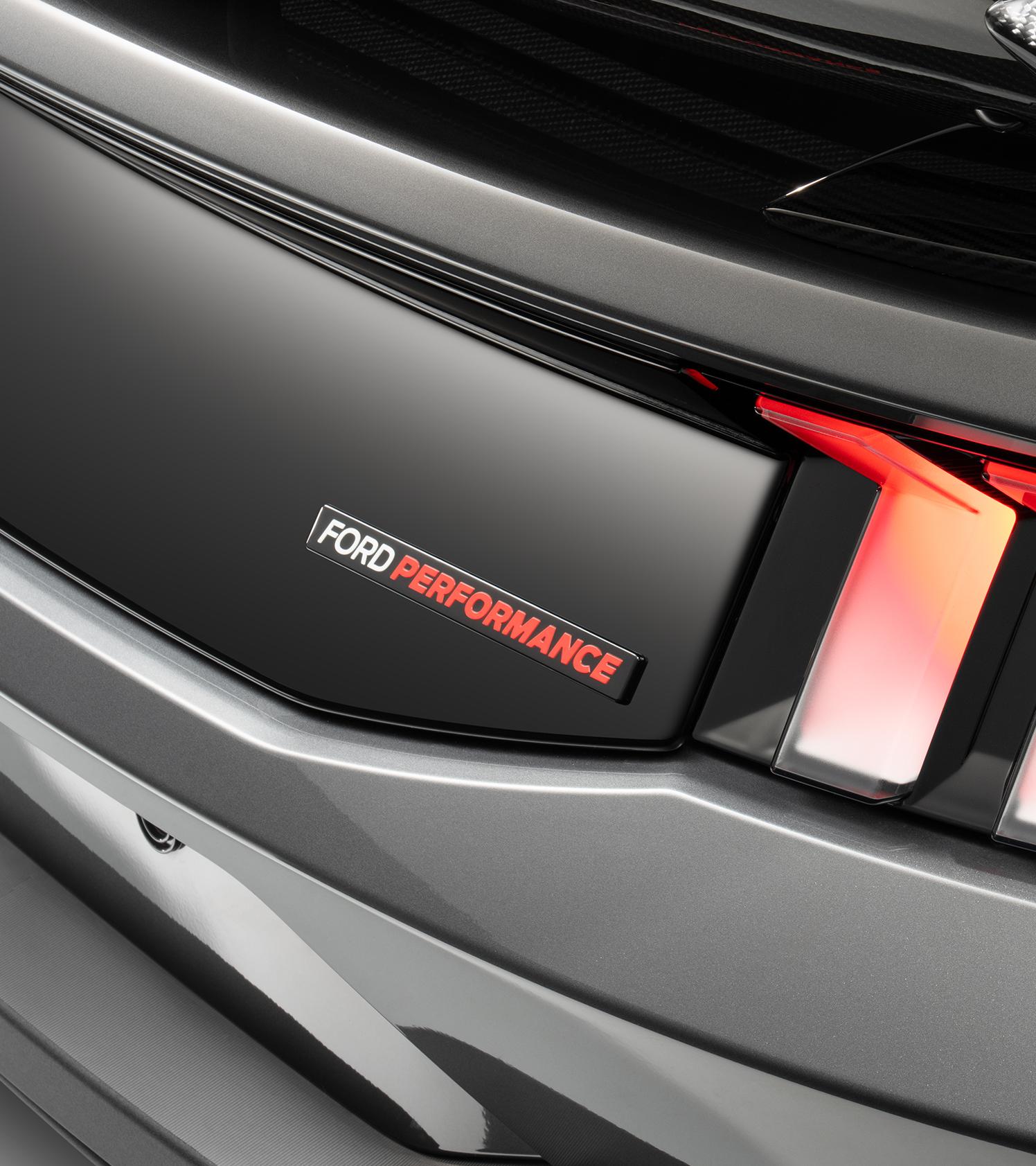 Ford Performance rear badge in white and red lettering on the gloss black decklid panel, next to the illuminated taillight of a gray Mustang