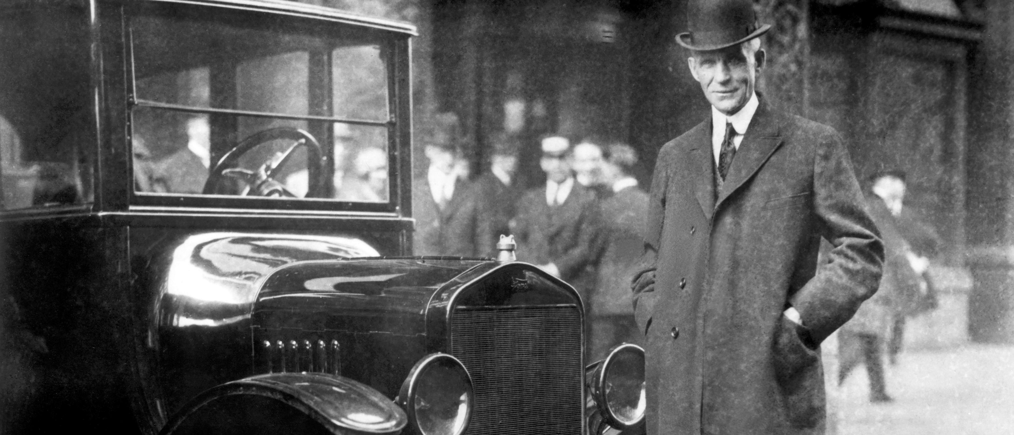 Henry Ford dressed in a bowler hat and overcoat stands next to a Ford Model T