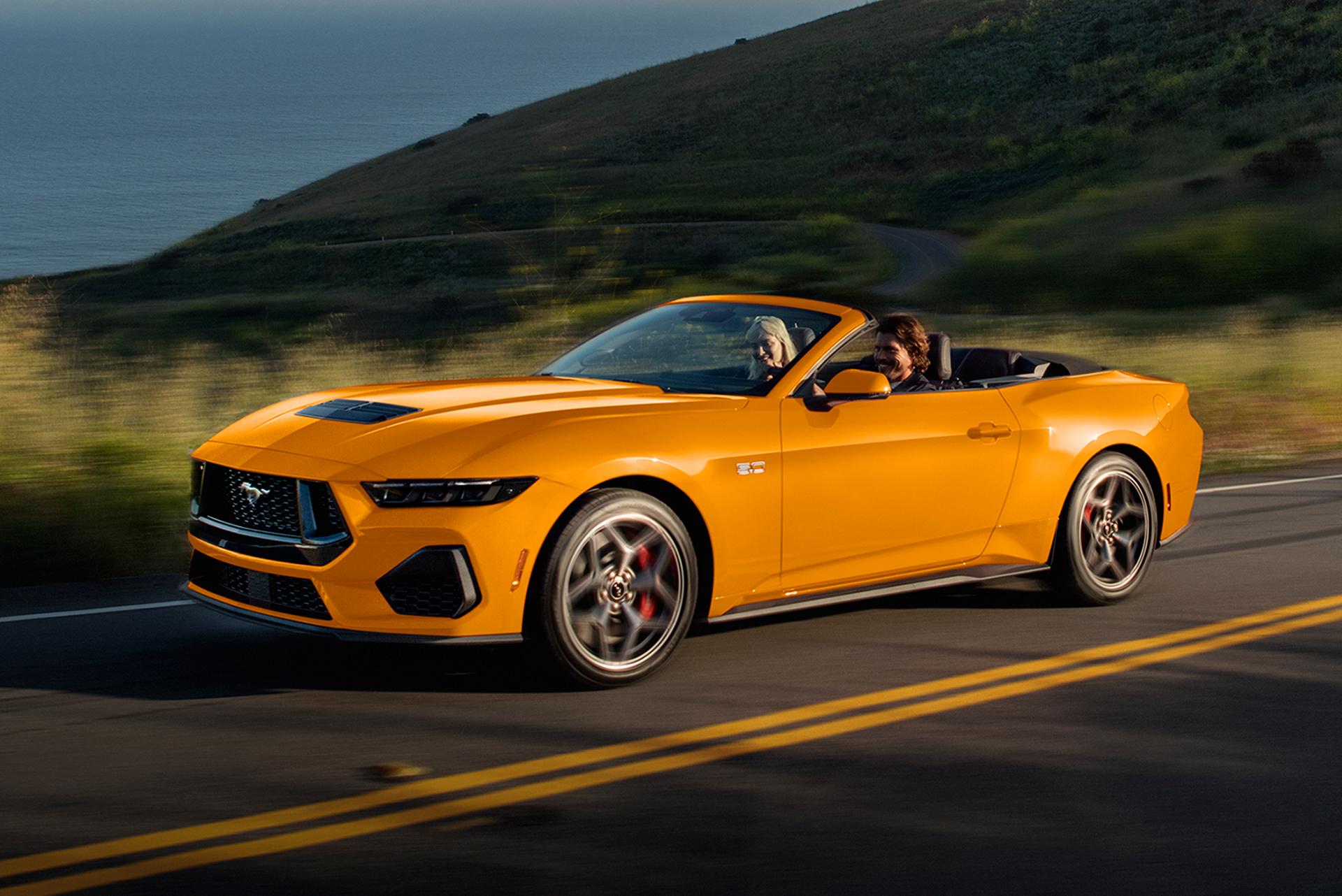Two people in a 2026 Ford Mustang® GT Convertible in Orange Fury Metallic Tri-coat being driven on a coastal highway