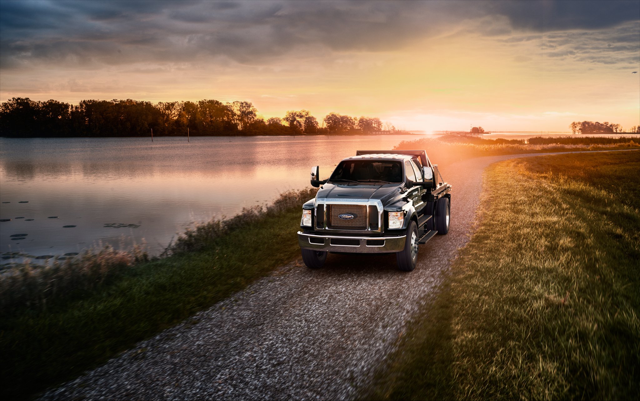 Ford F-750 2025 à cabine 6 places aménagé avec plateau à poteaux utilitaires en noir agate se déplaçant sur une route de gravier au coucher du soleil