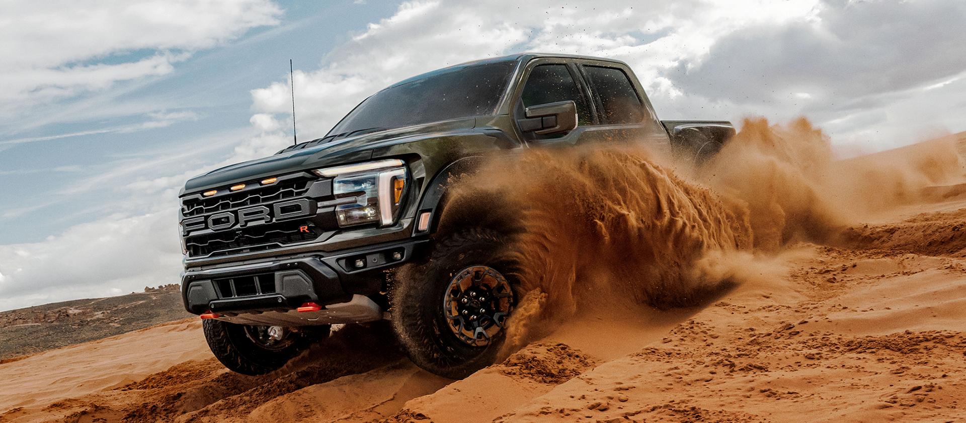 A black 2025 Ford F-150® Raptor® sends a wave of sand flying as it carves through a desert dune under a partly cloudy sky