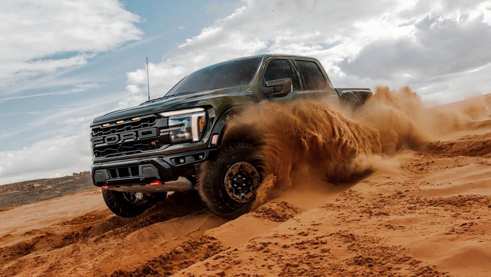 A black 2025 Ford F-150® Raptor® sends a wave of sand flying as it carves through a desert dune under a partly cloudy sky