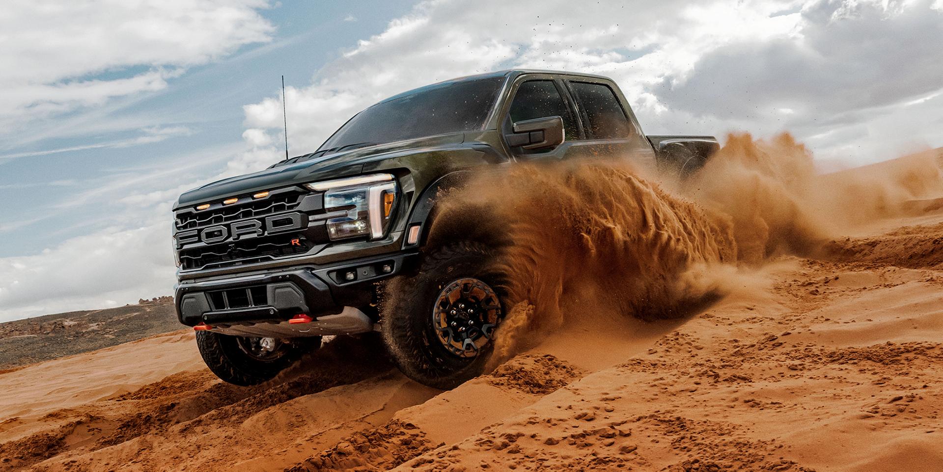A black 2025 Ford F-150® Raptor® sends a wave of sand flying as it carves through a desert dune under a partly cloudy sky
