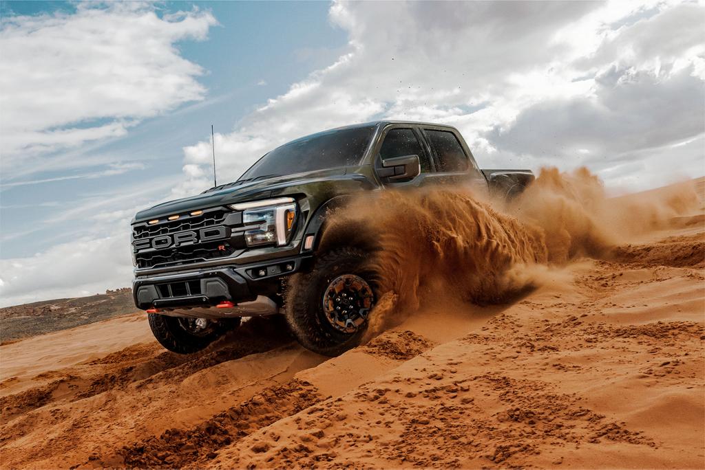 A black 2025 Ford F-150® Raptor® sends a wave of sand flying as it carves through a desert dune under a partly cloudy sky