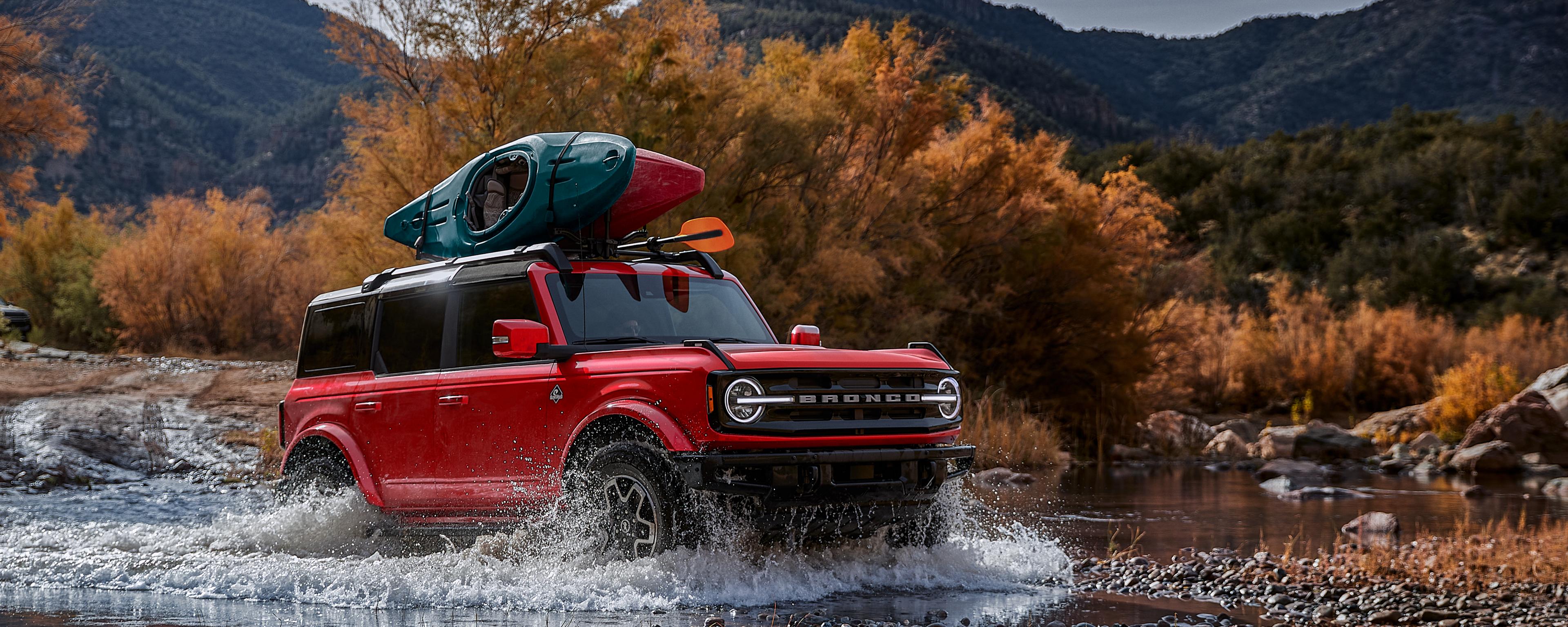 A 2025 red Ford Bronco® SUV splashing through a river with two canoes on the roof rack