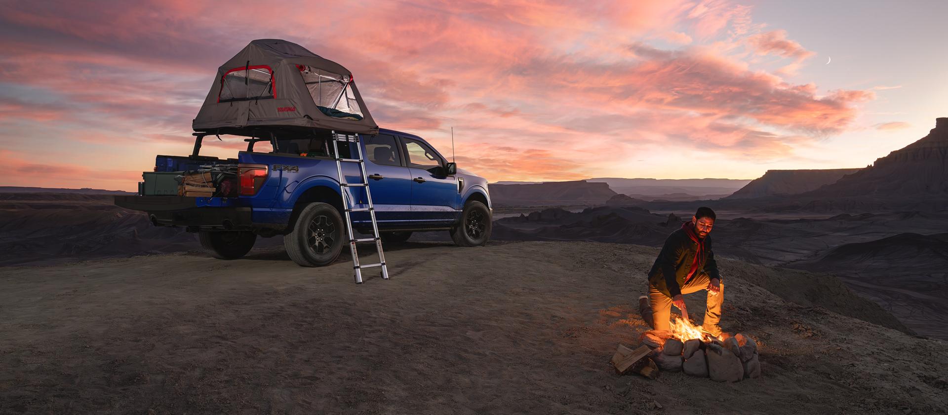 Man starting a campfire near a parked 2026 Ford F-150® STX® FX4 model with a Ford Accessories tent at dusk