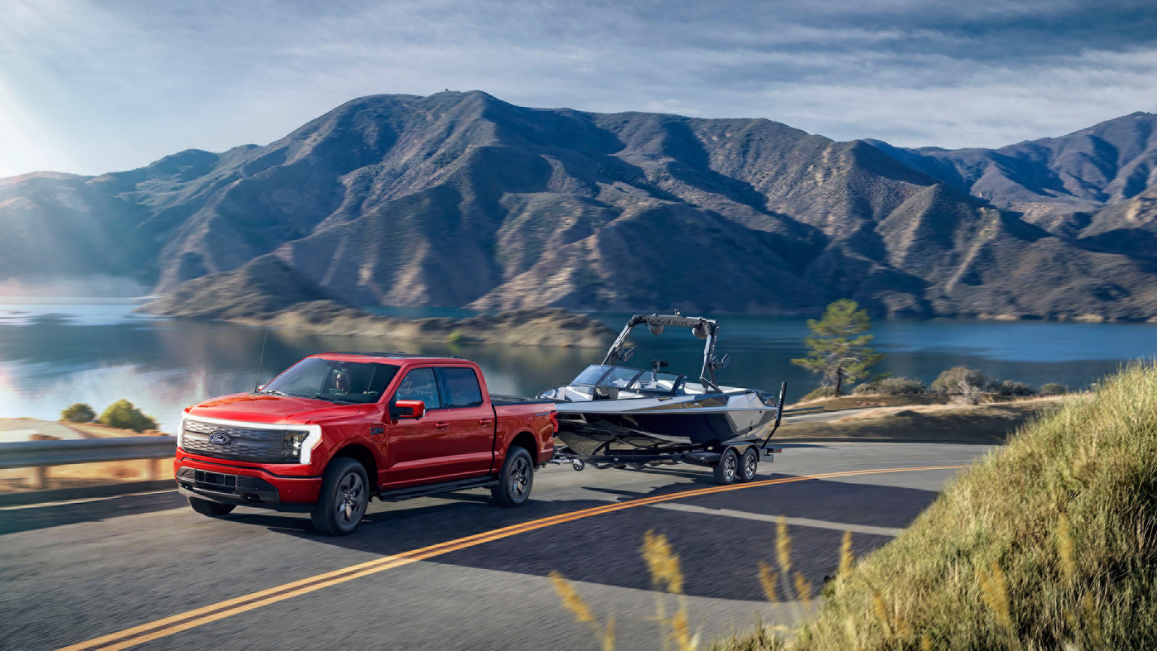 A 2025 Ford F-150® Lightning® pickup pulling a boat on a trailer down a highway near a mountain lake