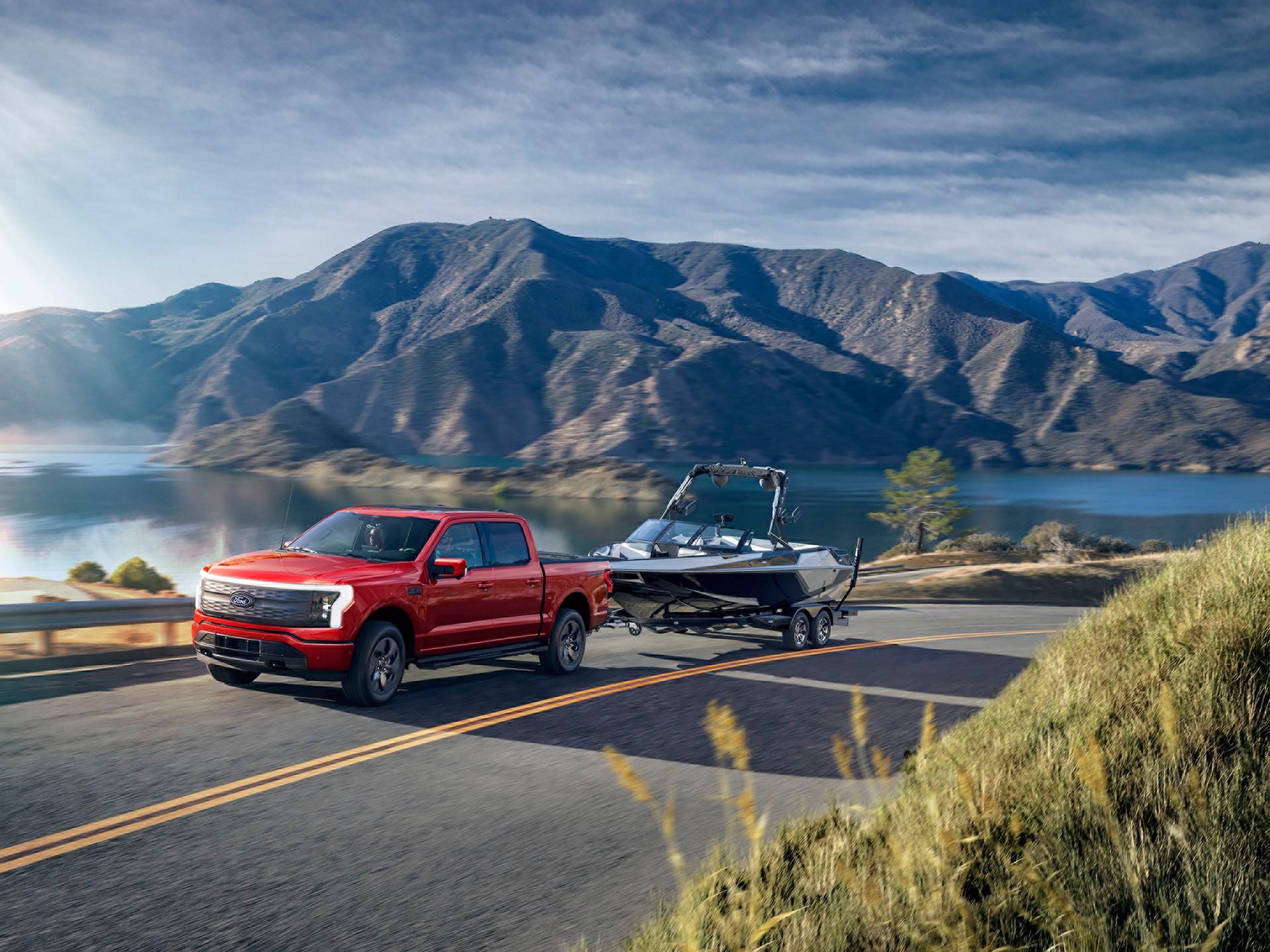 A 2025 Ford F-150® Lightning® pickup pulling a boat on a trailer down a highway near a mountain lake