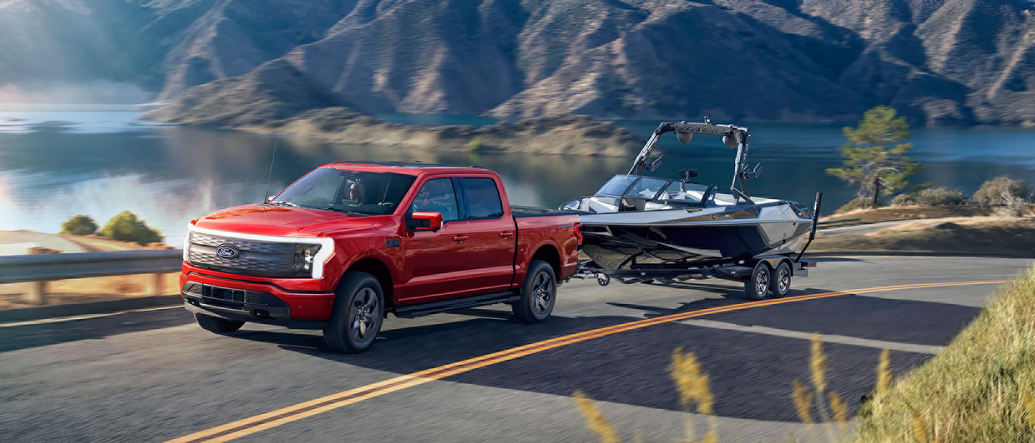 A 2025 Ford F-150® Lightning® pickup pulling a boat on a trailer down a highway near a mountain lake