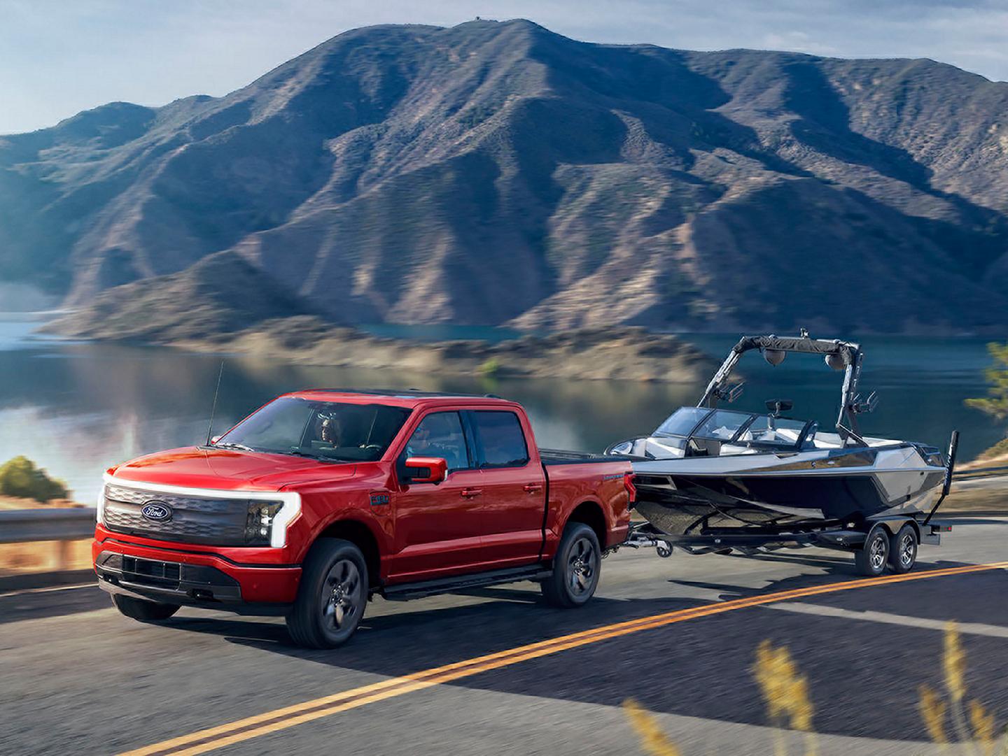 A 2025 Ford F-150® Lightning® pickup pulling a boat on a trailer down a highway near a mountain lake
