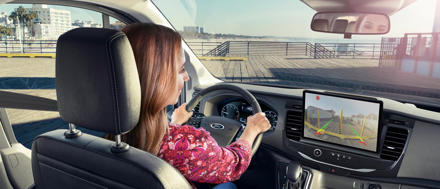 A woman driving a 2025 Ford Transit® van looking at her touchscreen seeing behind the van