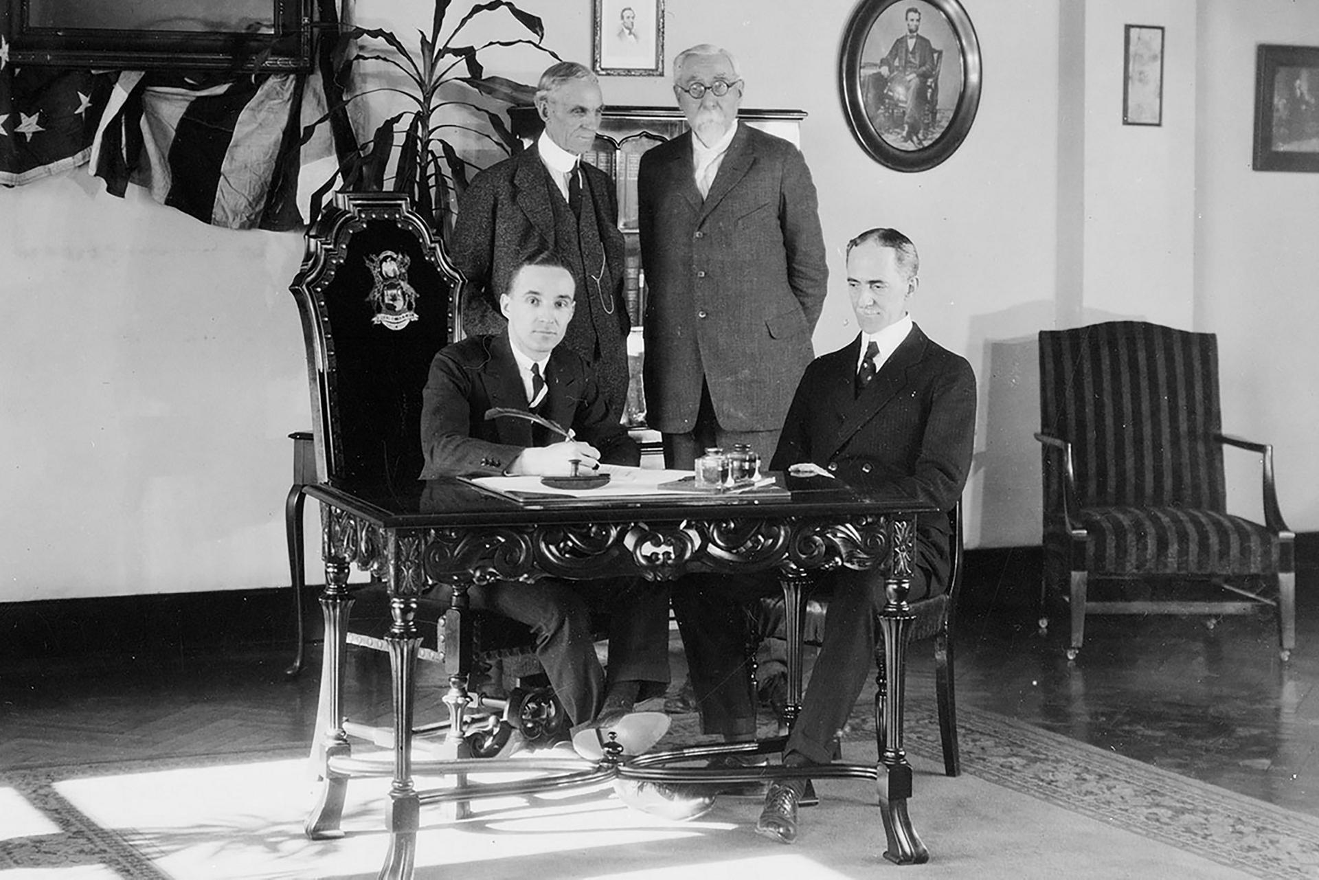 A black and white image of four men standing around a desk