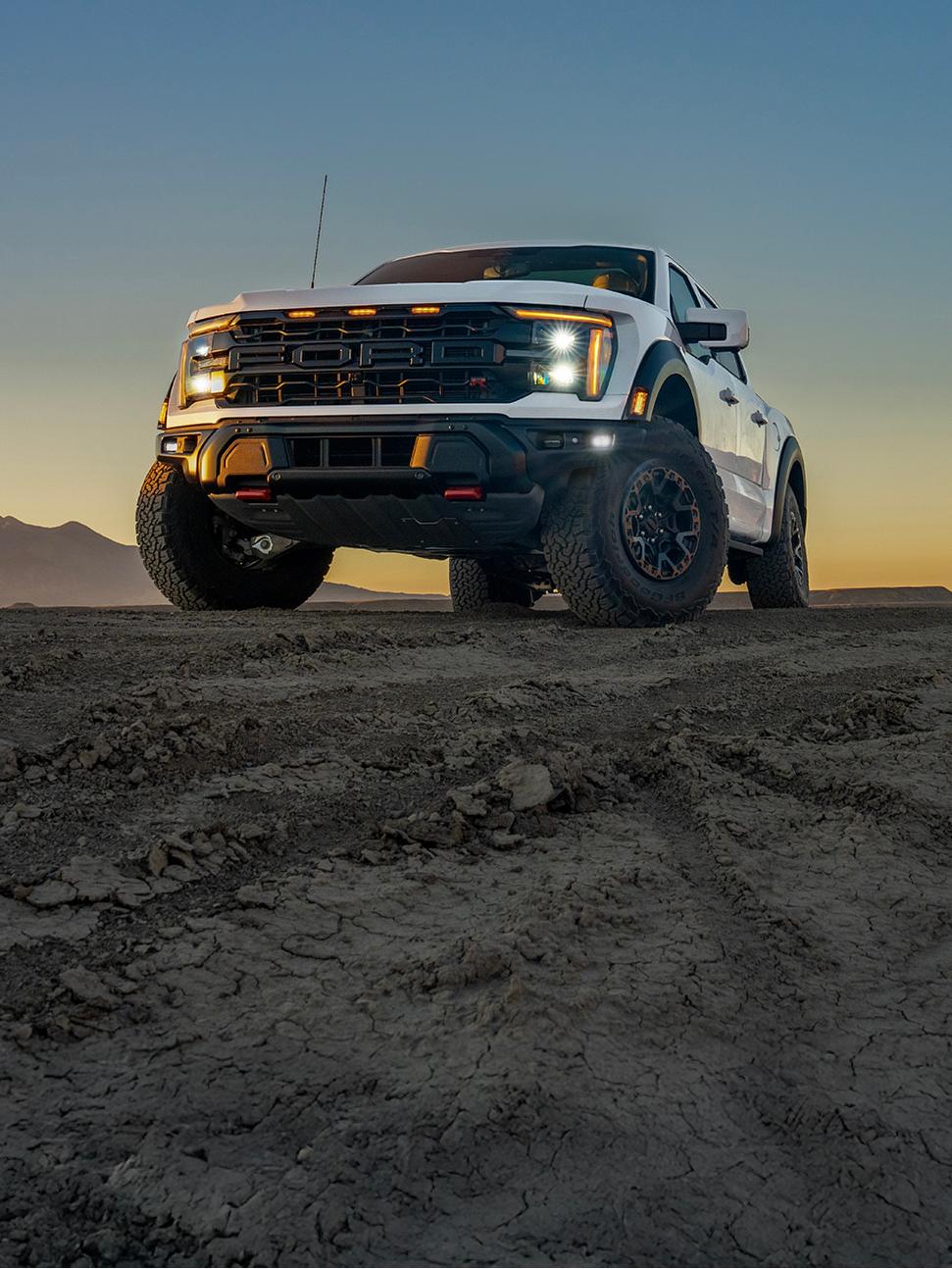 A white 2025 Ford F-150® Raptor® R™ shown from a low angle on rocky terrain, with the sunset and mountains behind it