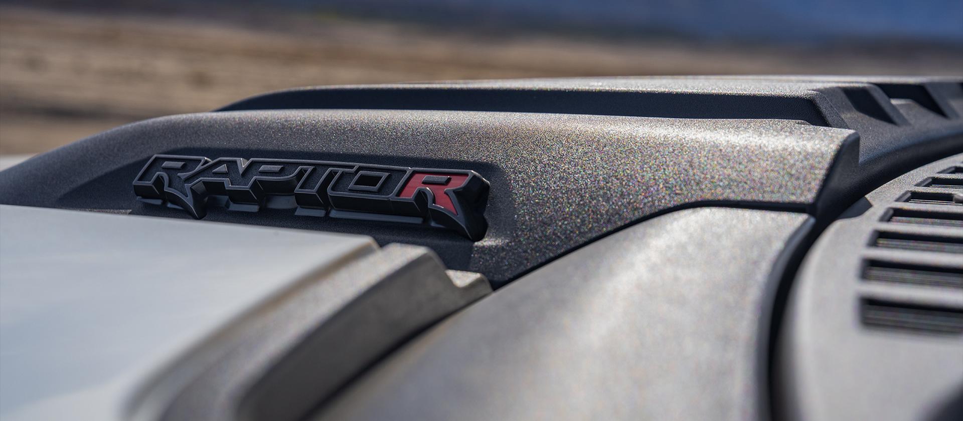 The front hood of a black 2024 Ford F-150® Raptor®, its distinctive red 'R' logo dimly lit by the hot desert sun