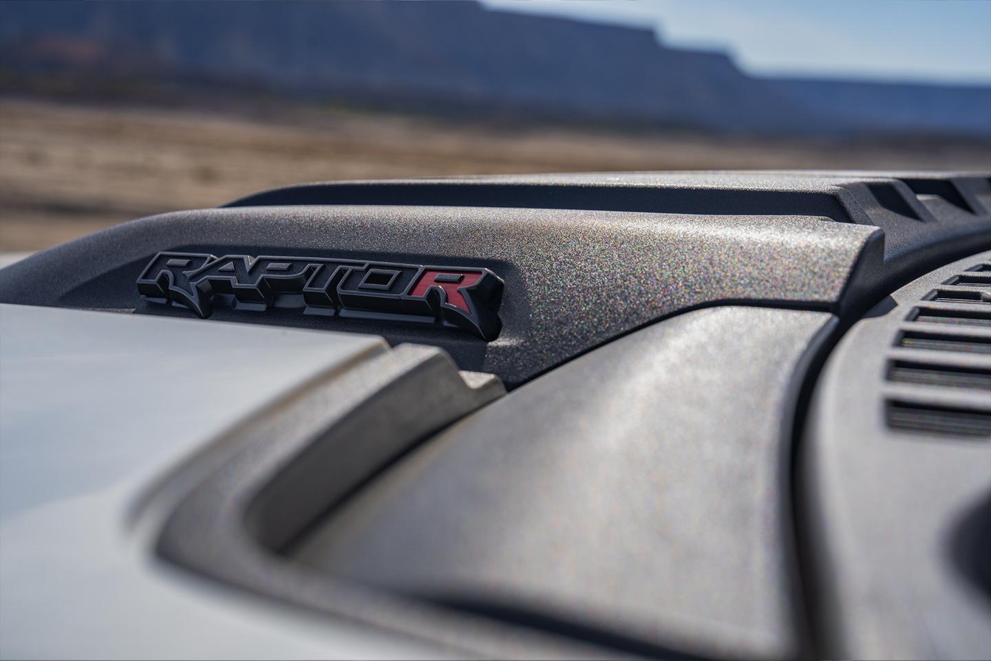 The front hood of a black 2024 Ford F-150® Raptor®, its distinctive red 'R' logo dimly lit by the hot desert sun