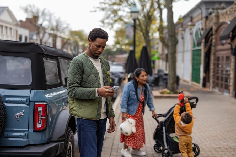 A family stands on a sidewalk beside their parallel-parked Ford Bronco® while the father looks at his phone