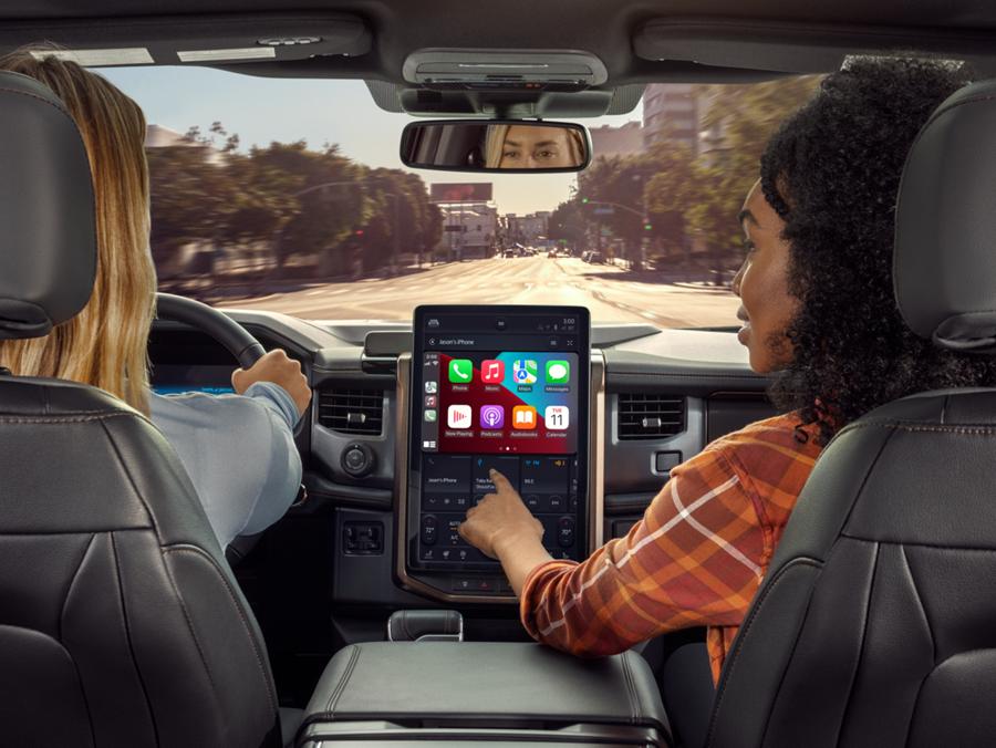 A view from the backseat of a Ford F-150 Lightning pickup truck, looking toward the front. A woman in the passenger seat is using her hand to manipulate the SYNC touchscreen, which is displaying the Apple CarPlay user interface. The woman driving has her hands safely on the wheel.
