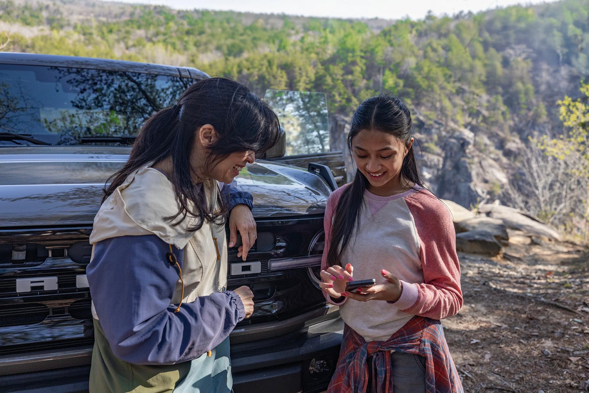 Two people laugh and smile at their phone while standing in front of a 2026 Ford Bronco SUV® parked in the mountains