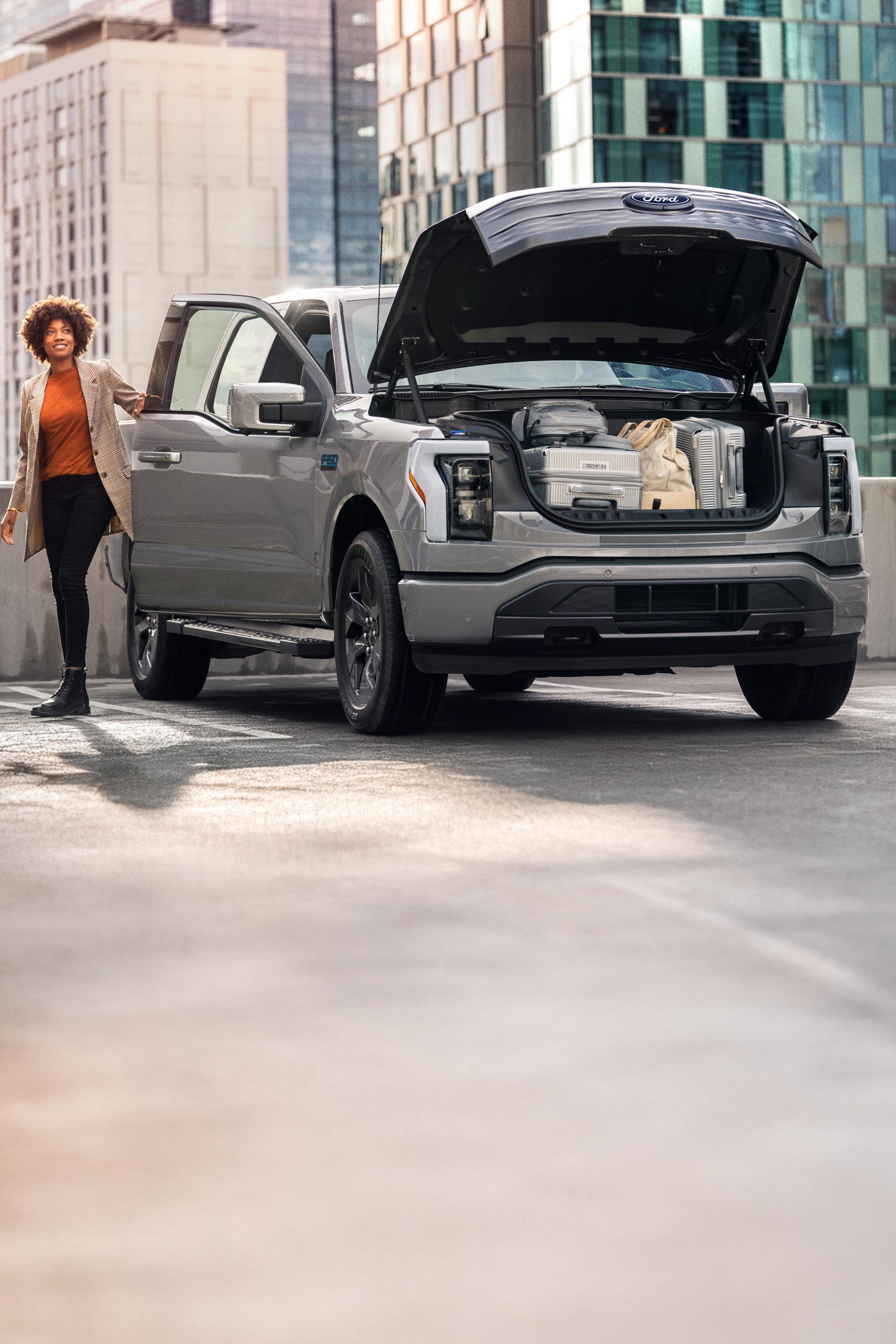 A gray 2025 Ford F-150® Lightning® truck parked with the frunk open. There are suitcases and bags in the frunk. A woman is on the passenger side heading toward the front.