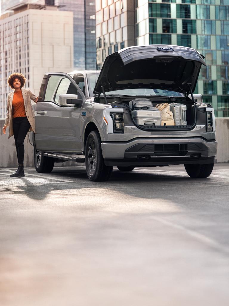 A gray 2025 Ford F-150® Lightning® truck parked with the frunk open. There are suitcases and bags in the frunk. A woman is on the passenger side heading toward the front.