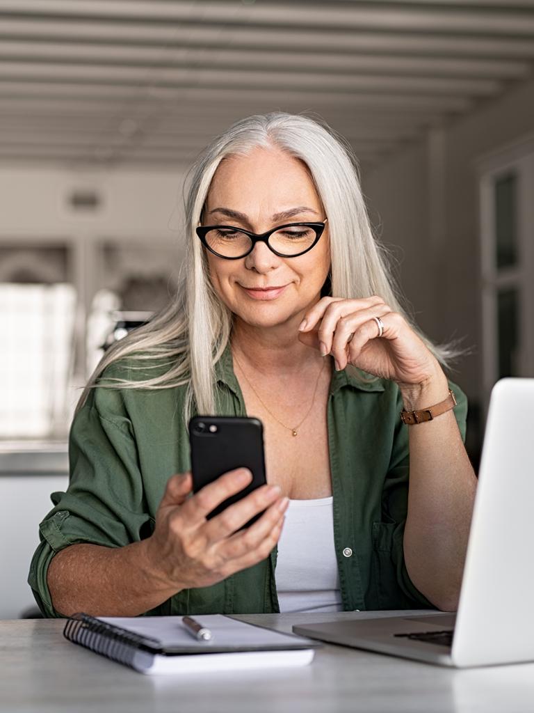 A person seated at a table in front of a laptop looks down at a phone in their hand and smiles