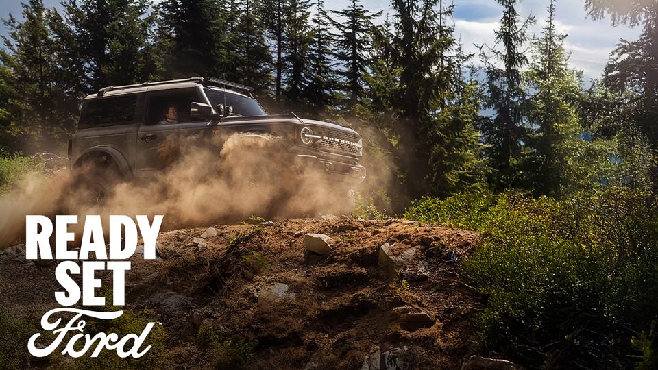 A 2026 Ford Bronco® SUV being driven on a muddy forest trail, with the legend “Ready Set Ford” superimposed