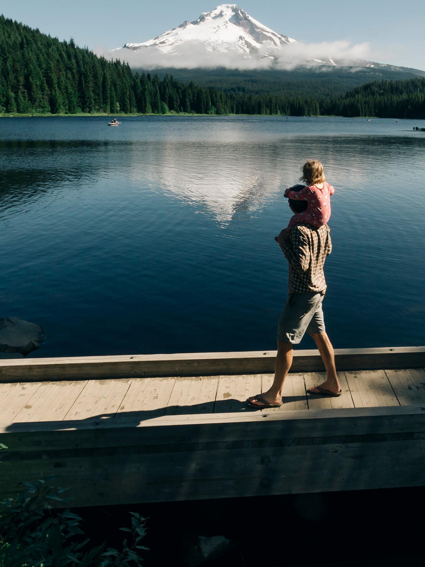 A Parent with a child on their shoulders walks beside a lake
