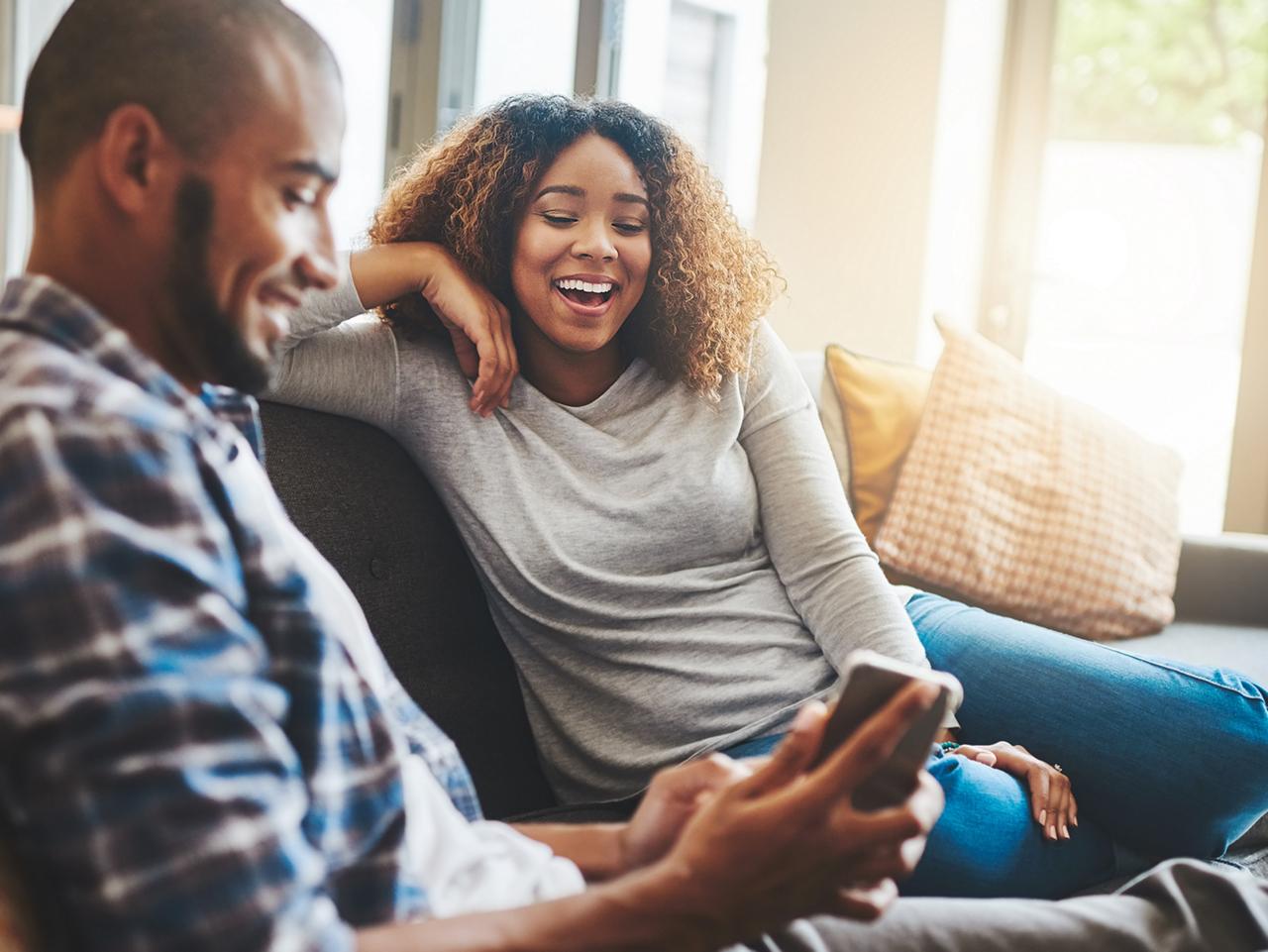 A couple sitting on a couch at home, smiling and looking at a smartphone together