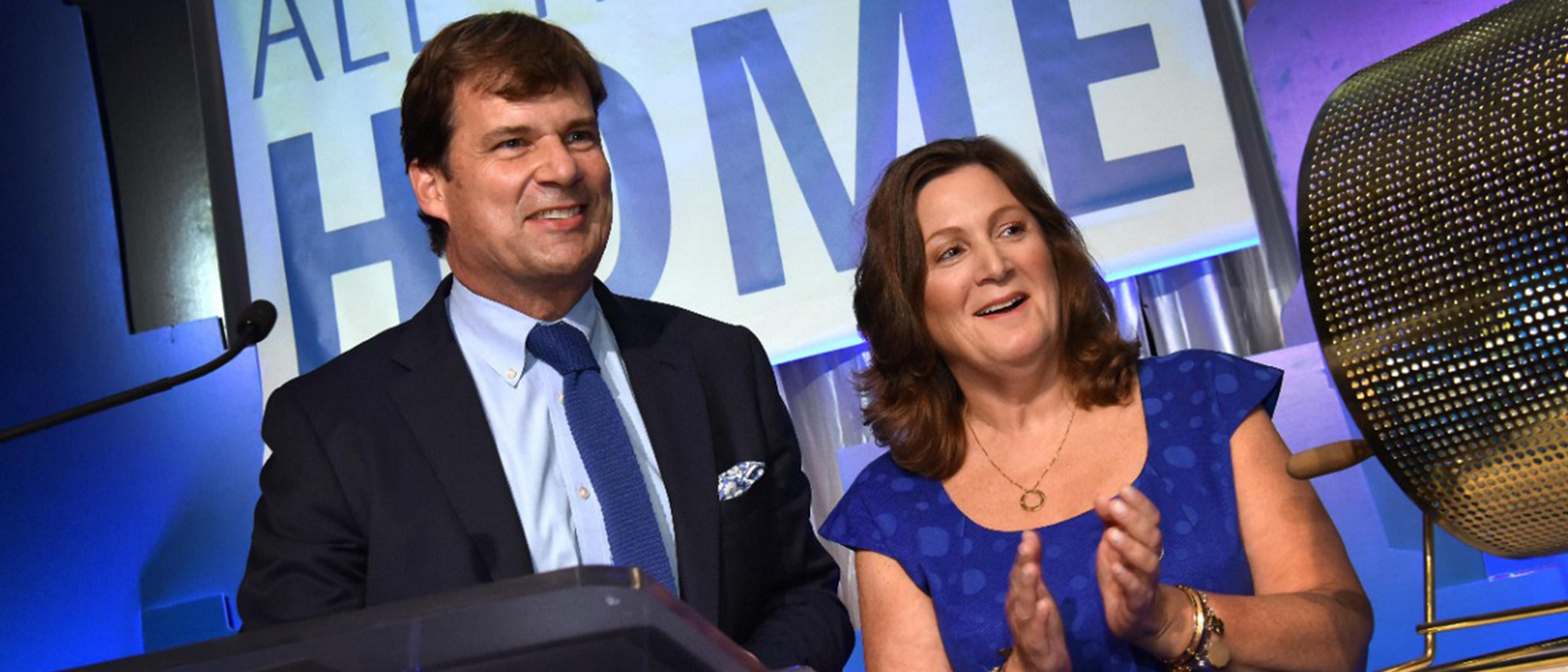Jim Farley and Elena Ford stand behind a podium at an event smiling and applauding the audience