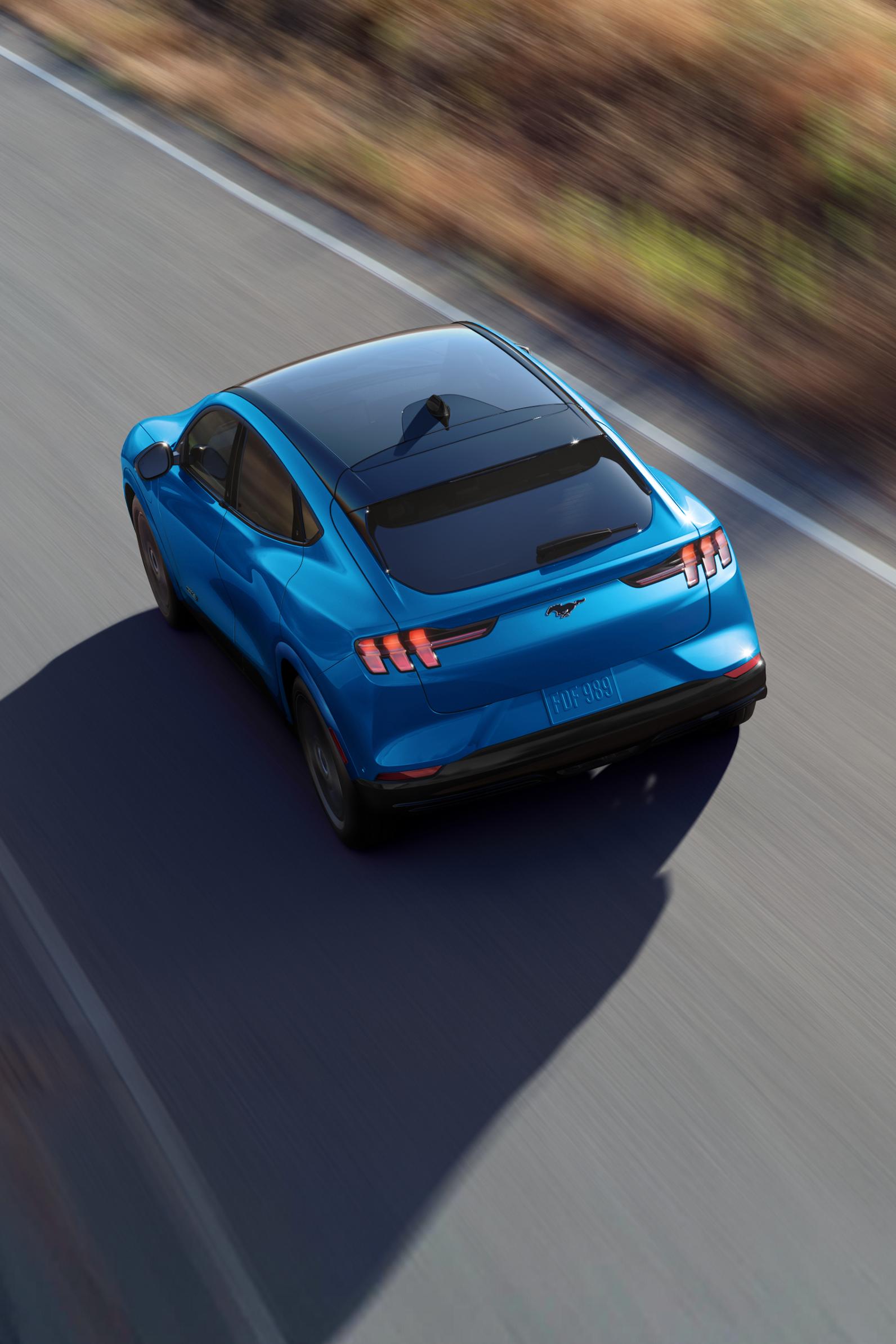 A blue 2025 Ford Mustang Mach-E driving along a paved road next to a field