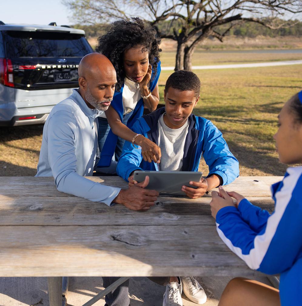A family sits at a picnic table looking at a tablet, using the Wi-Fi from the Ford Expedition parked in the background.
