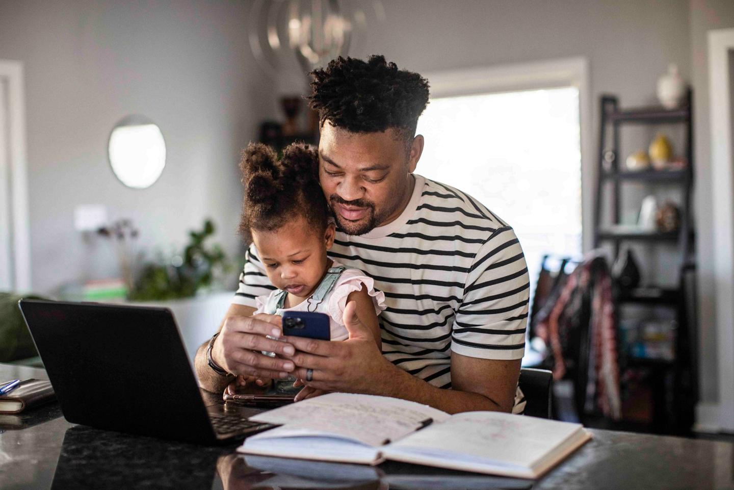 A man uses his smartphone, while his daughter sits on his lap and looks on interestedly