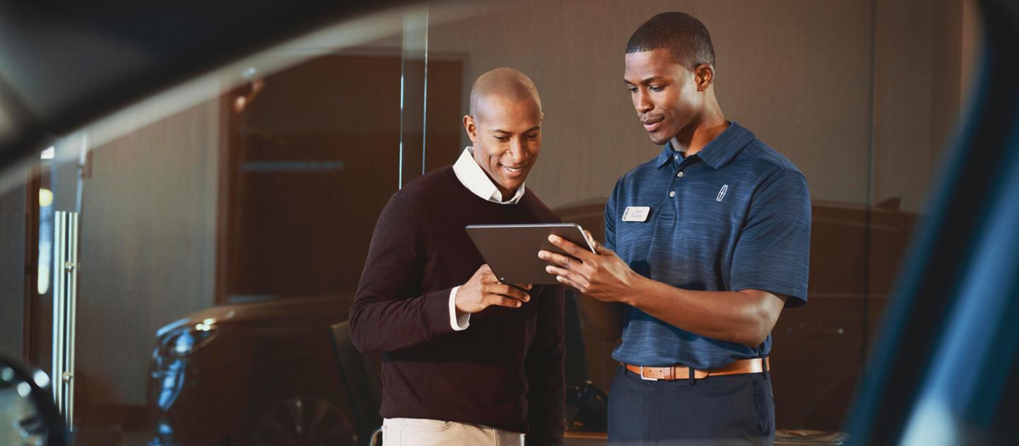 A man wearing a nametag and a Lincoln logo polo shirt shows information on a tablet computer to another man, as seen through the window of a Lincoln SUV