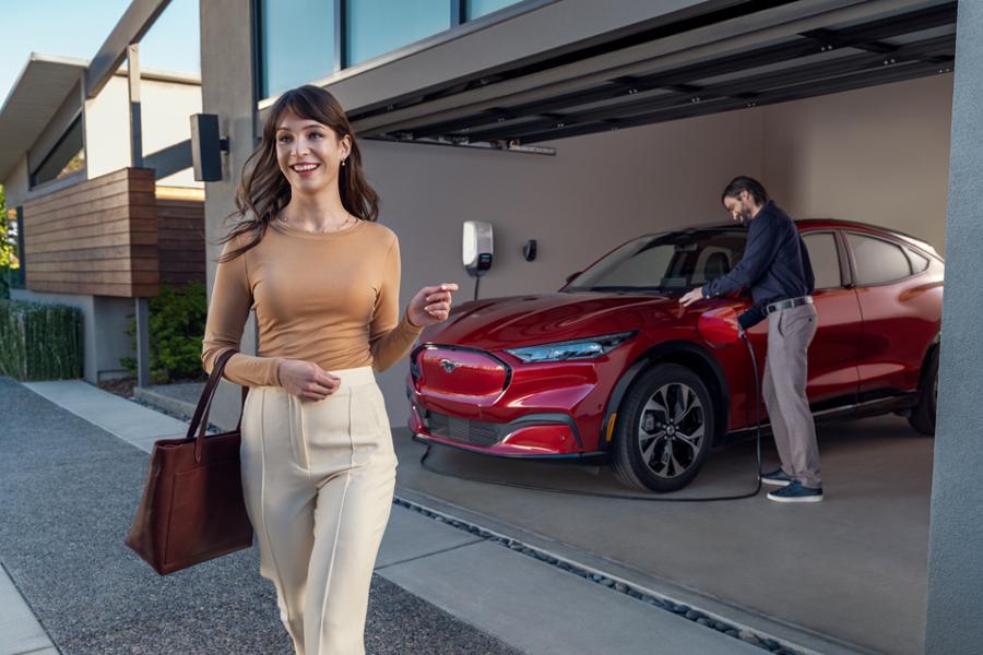 A woman walking away from a red Ford Mustang Mach-E SUV parked in a garage, while a man plugs a charging cable into the vehicle's port