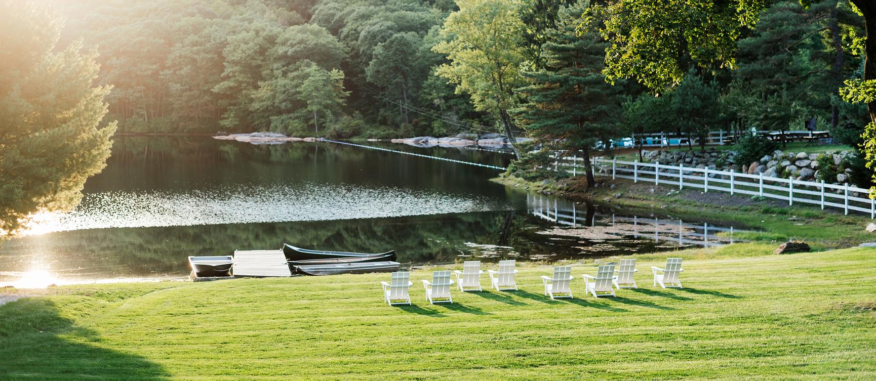 A scene of a green lawn featuring white deck chairs placed around the lake’s edge at Cedar Lakes Estate, a luxury retreat set on a former summer camp.