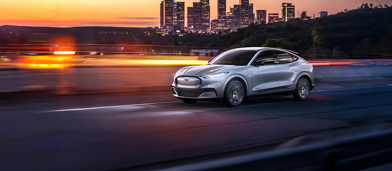 2025 Ford Mustang Mach-E driving down road at sunset with city in background