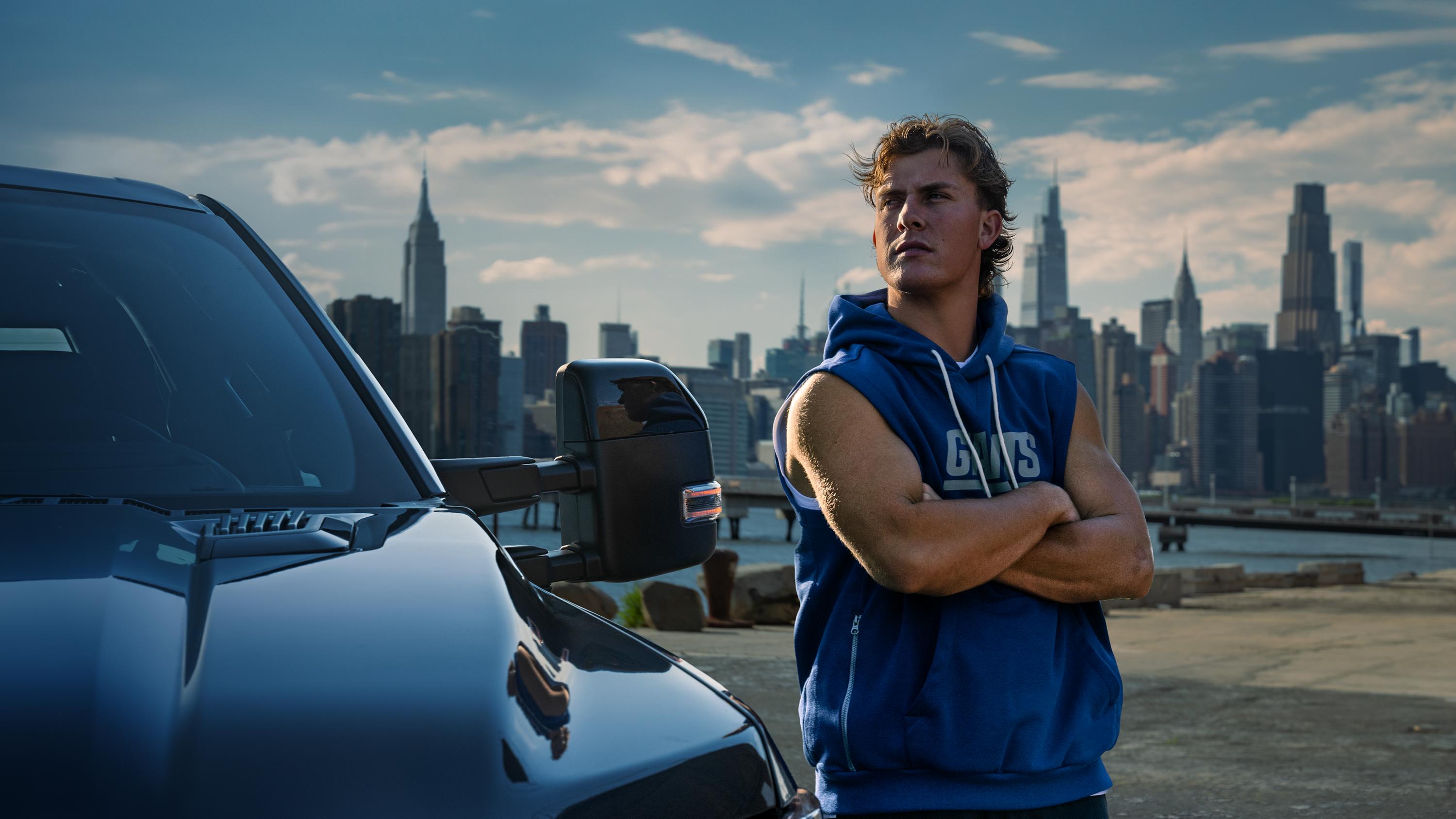 A silver Ford F-150® Truck is parked in front of the New York City skyline. New York Giants Quarterback Jaxson Dart stands proudly by the truck.