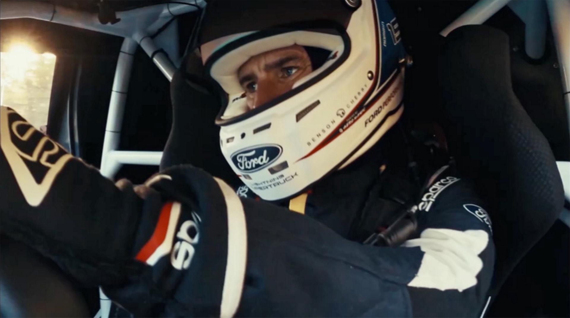 Ford race team member shown driving wearing flight suit and racing helmet