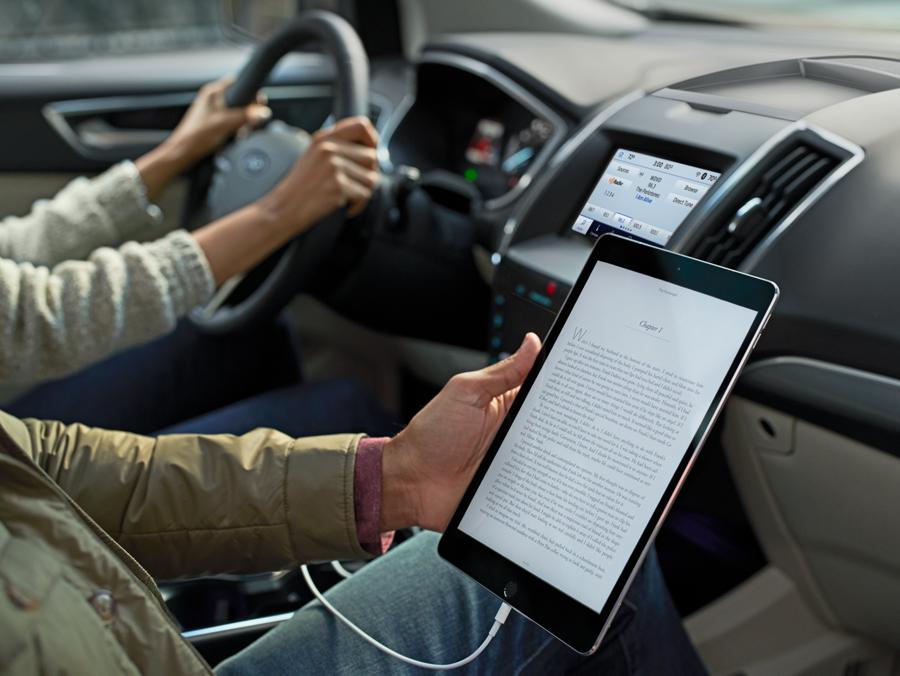 A man in the passenger seat of a 2020 Ford Edge reads an e-book on his tablet computer, which is connected to the vehicle with a USB cable. In the background, the vehicle's driver keeps her hands safely on the wheel.