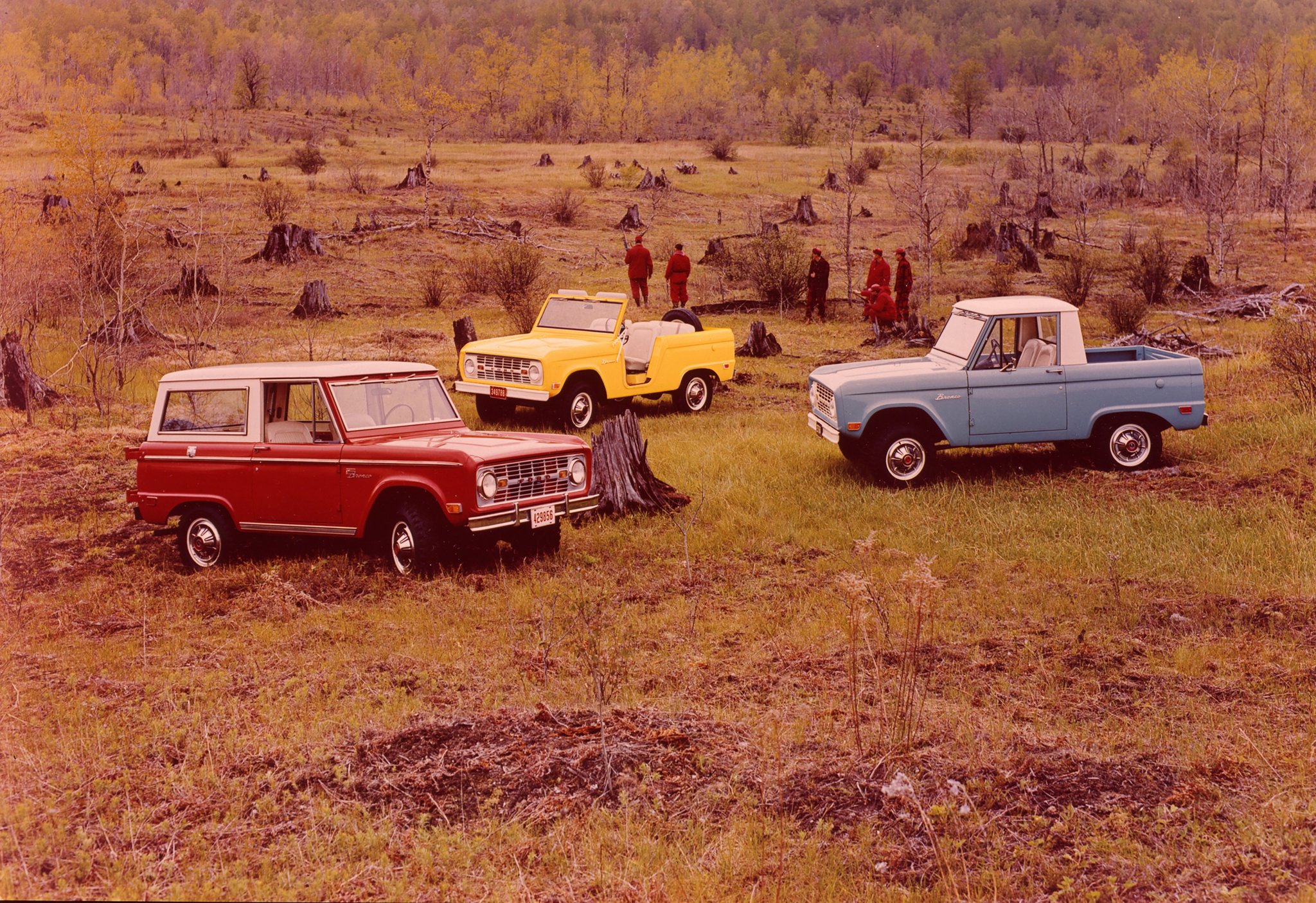 Three 1969 Ford Broncos parked in a wilderness field while a group of people walk nearby