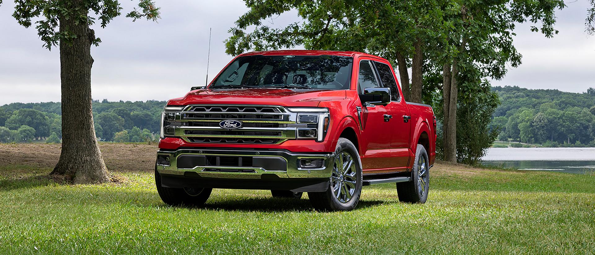 A red 2027 Ford F-150® Lariat® pickup parked in a tree-lined field with a lake scene behind it