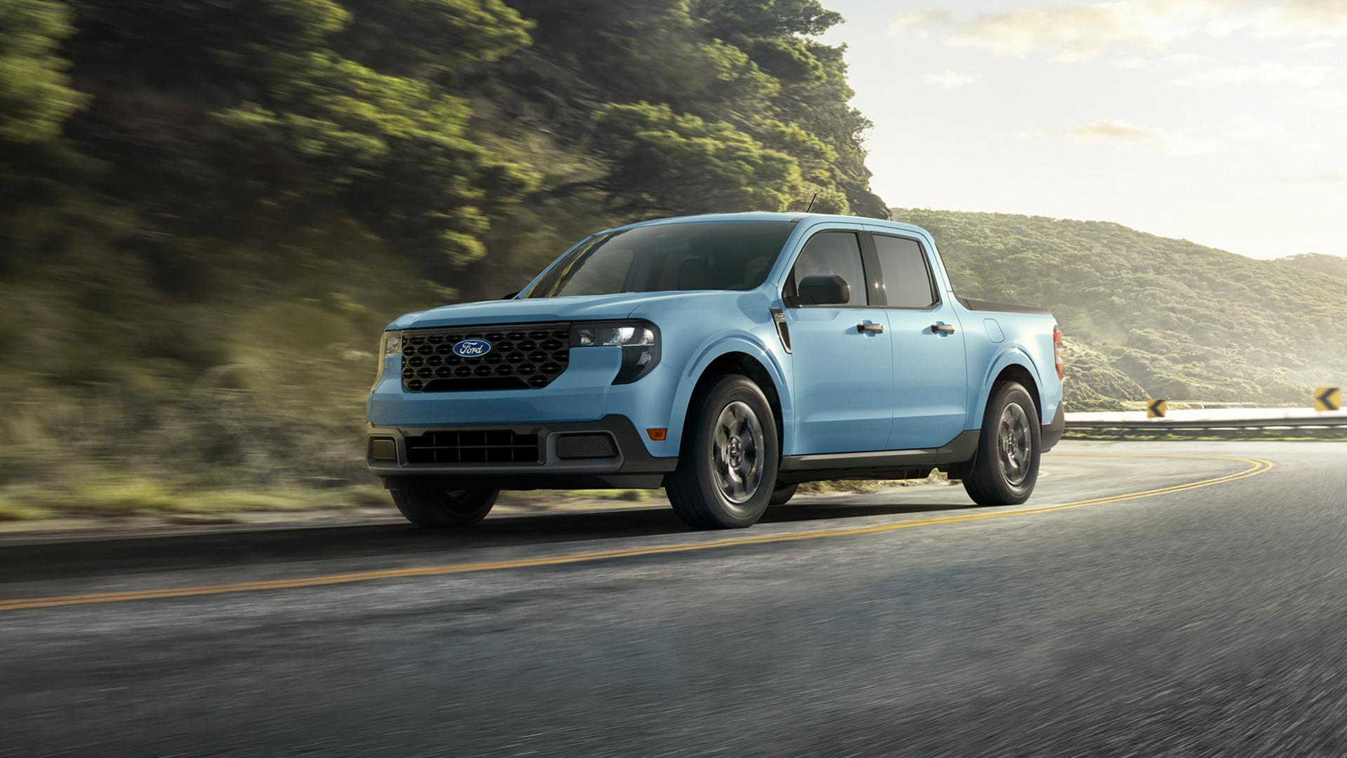 A gray 2026 Ford Maverick® pickup driving down a winding highway near a mountain lake