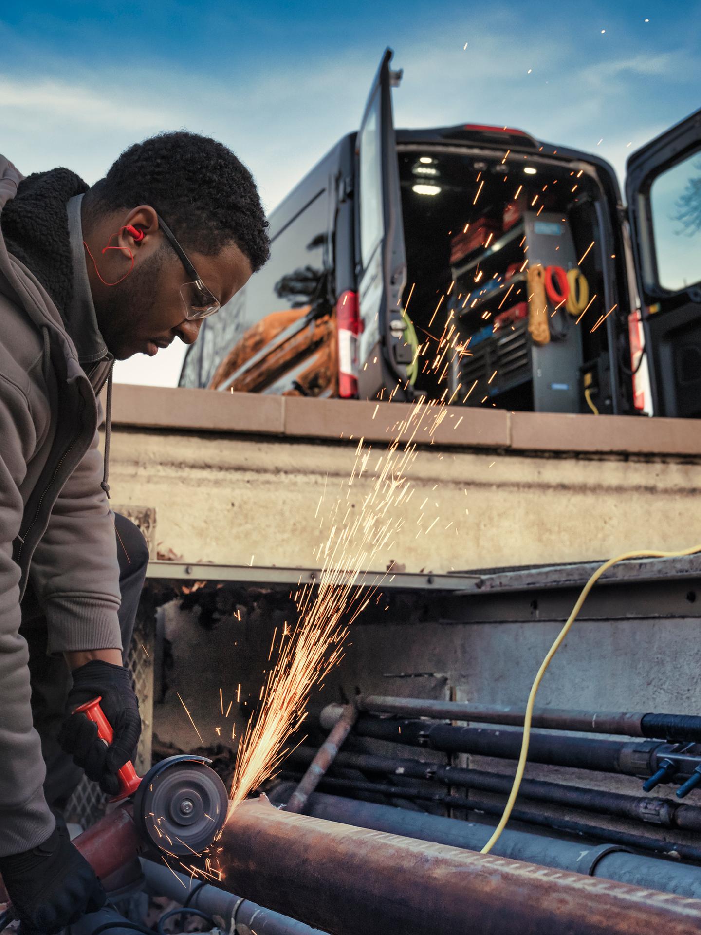 A worker creating sparks with a power tool near the open rear doors of a dark 2027 Ford Transit® Van