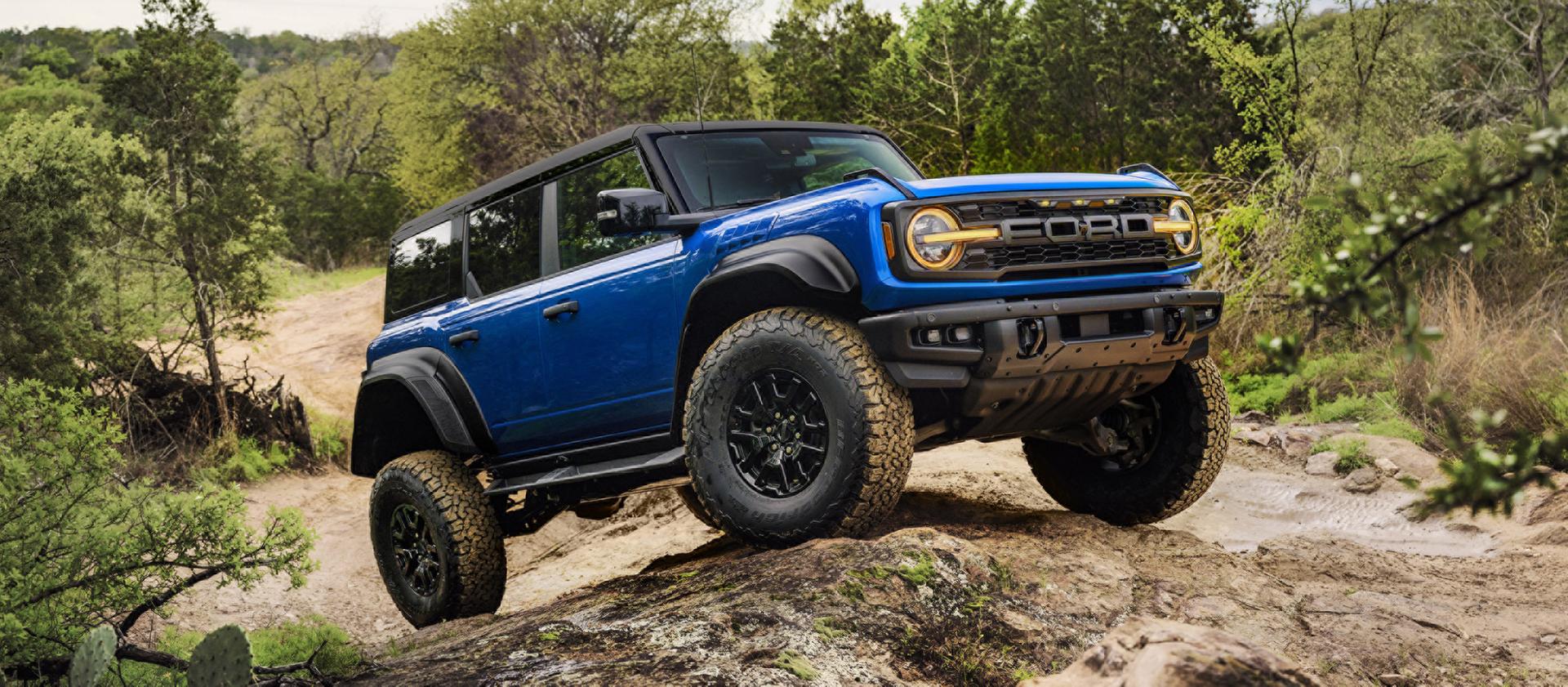 A blue 2025 Ford Bronco® Raptor® climbing over a large rock on a wooded trail, showing off its suspension articulation