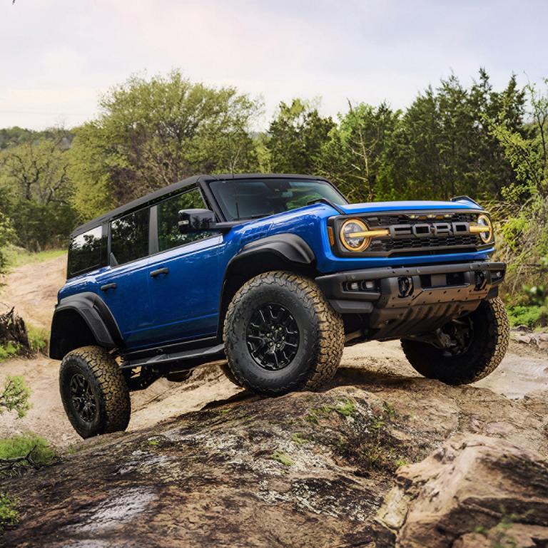 A blue 2025 Ford Bronco® Raptor® climbing over a large rock on a wooded trail, showing off its suspension articulation