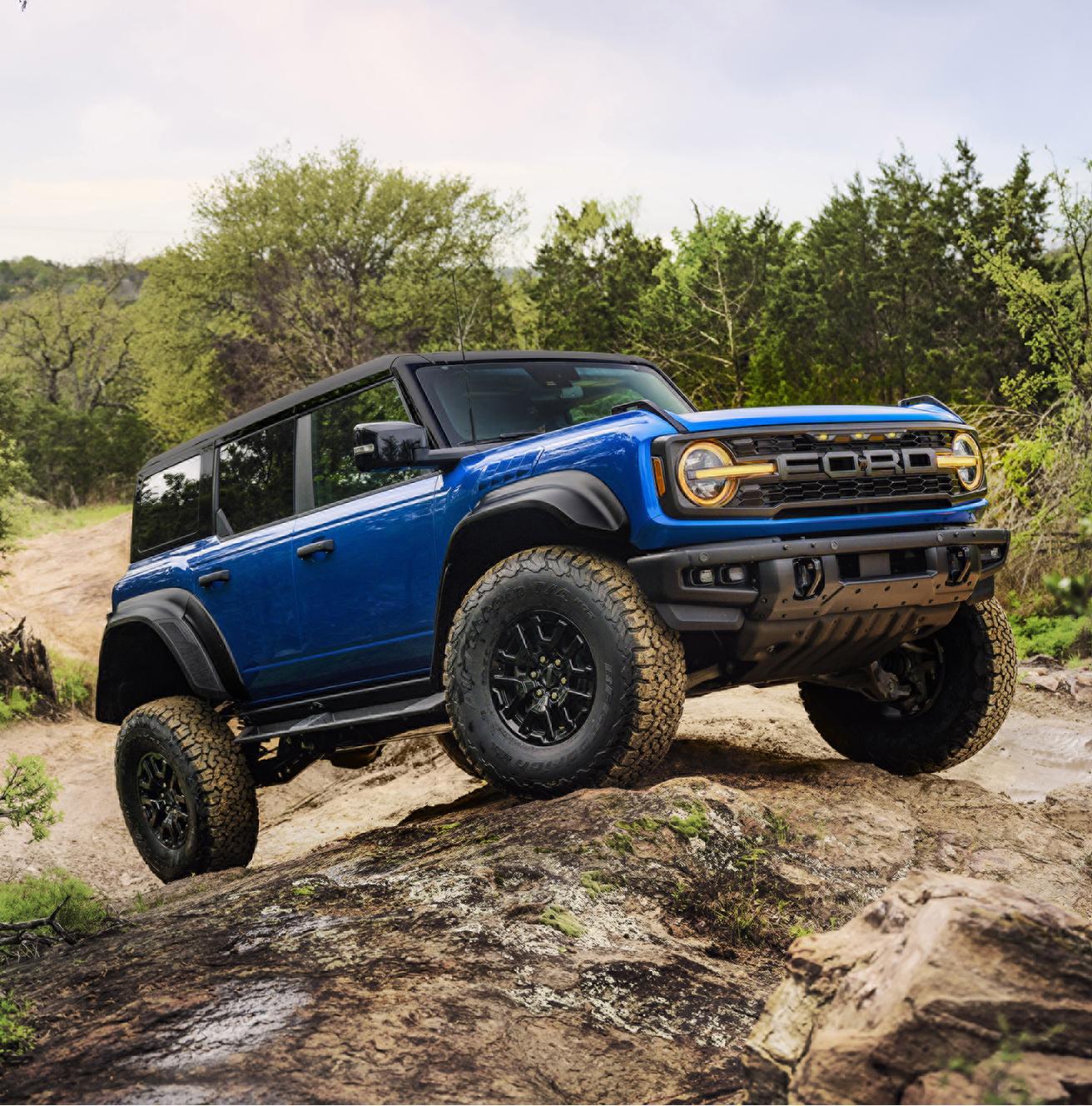 A blue 2025 Ford Bronco® Raptor® climbing over a large rock on a wooded trail, showing off its suspension articulation